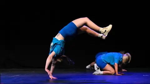 A woman wearing blue t-shirt and shorts does a back flip on stage while another person is on their hands and knees in the background.