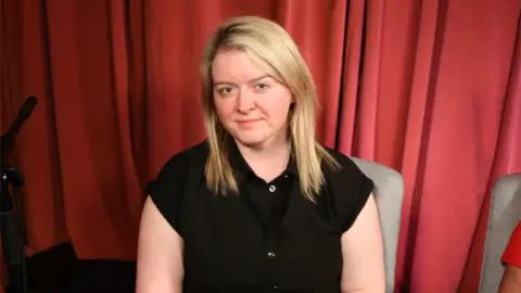 A woman with blonde shoulder-length hair and a black shirt sits on a chair, in front of a red curtain.