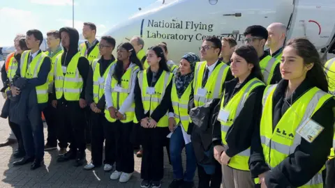 Students from Luton wearing high-vis jackets pose for a photo in front of an aircraft - the National Flying Laboratory Centre. 