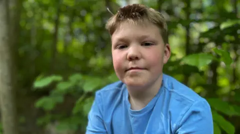 A boy, in woodland is pictured looking at the camera. He has blonde hair and is wearing a blue t-shirt