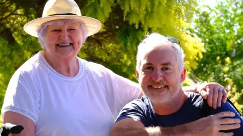 Beryl and Peter smile at the camera. Beryl has short white hair and is wearing a large sun hat and has her arm around the shoulder of Peter, who has short white hair and a white beard. Behind them are green trees. 
