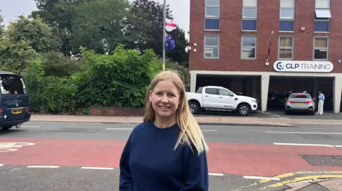 BBC A blonde wearing a blue crew-necked jumper stood alongside a road with a lamppost in the background with an England and an Australian flag flying