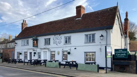 Luke Deal/BBC The front of the two-storey Kings Head pub. It is painted white, with brown tiles on the roof. The pub sign is of Henry VIII. Picnic tables are out the front next to the pavement.