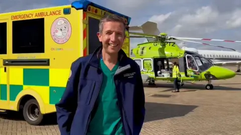 A smiling man in medical scrubs stood in front of an ambulance and helicopter