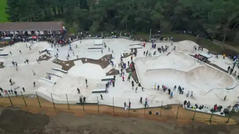 A picture taken from above looking down on Les Quennevais skate park. It shows white skate bowls and people gathered around them. 