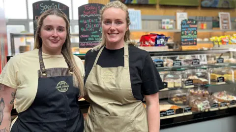 Lily and Honey Kennedy smile. A range of cakes are displayed in a counter behind them. They both have long straight blonde hair and wear it half up half down. Lilly is wearing a butter yellow t-shirt and a black apron. Honey is wearing a black t-shirt and a butter yellow apron. Both aprons have Cafe Cenno's logo.