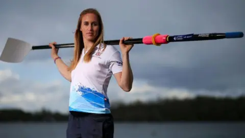 Nick Potts/PA Wire Helen Glover, standing outdoors near a body of water, holding a rowing oar across her shoulders. She has long, light brown hair and is dressed in sportswear, including a white top with blue accents and branding. The background is an overcast sky with distant trees.