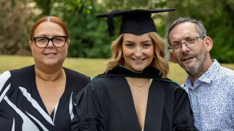 NHS Humber Health Partnership A woman with curled blonde shoulder length hair is wearing a light yellow dress and gold necklace underneath a black graduation gown and cap. She is smiling next to a woman with red hair and black glasses on and black and white top, and a man with black rectangular glasses and a white patterned shirt.
