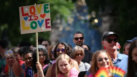 Getty Images A member of the public watching the parade holds a placard with rainbow letters saying "Love is love". They are surrounded by other people all looking in the same direction