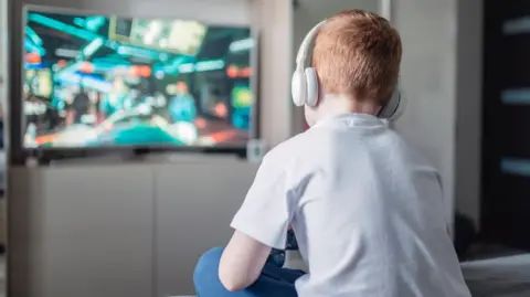 A boy with ginger hair is sitting down with his back to the camera, playing a video game on a large tv in the background. He has white headphones on and is wearing a white t-shirt and blue jeans.