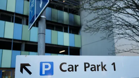 Shaun Whitmore/BBC Car Park 1 at Addenbrooke's Hospital in Cambridge. It is a multi-storey car park with blue and green panels on the exterior. On part of the outside there is smoke damage and the once blue and green panels are black following a fire. In the foreground of the picture there is a white sign with an arrow pointing to the left and writing which says, Car Park 1. 