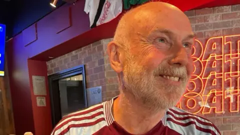 Neil Gilbert has a white beard and is wearing a claret and blue football shirt. He smiles as he looks to our right while sitting in a pub.