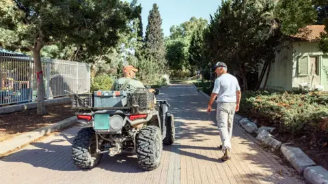 Maya Meshel / BBC Rami and Simon, some  with their backs to the camera. Rami is connected  an all-terrain vehicle. Simon is walking to the close    of him.