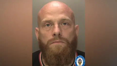 West Midlands Police A mugshot of a man with a ginger beard and bald head. He is wearing a dark t-shirt with a red and white collar.