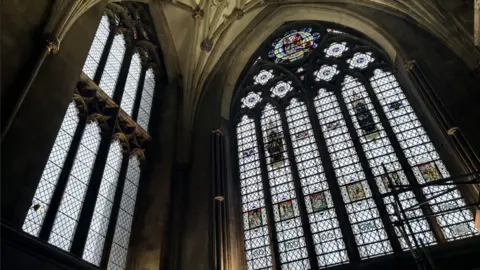 The inside of Bristol Cathedral showing the stained-glass Colston's window next to a plain window to its left. 