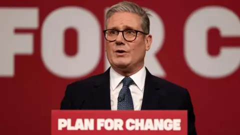 Starmer stands at a red lectern branded with the Labour Party slogan "Plan for Change", speaking as he looks slightly to the left