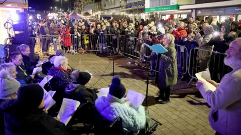 Karbon Homes A crowd of people behind metal barriers watching and taking pictures of the singers. Sara Dennis is at the front singing facing the singers with a blue folder in her hand. She has short, blonde hair and is wearing a green coat. In front of her is the older group of singers who are facing away from the camera. They are wearing coats and hats and holding the lyrics in their hands.