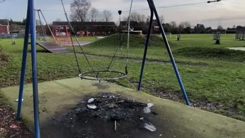 A circular swing is suspended from play equipment with no basket in the middle. The tarmac underneath it is burnt, scorched black with the remainder of the basket swing and the rest of the tarmac is green. A field and other play apparatus can be seen behind it.