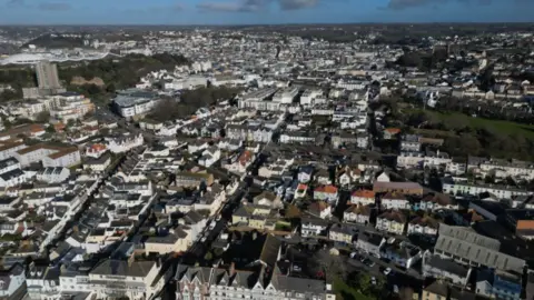 An ariel view of St Helier in Jersey. Dozens of rows of houses are visible under a blue sky.