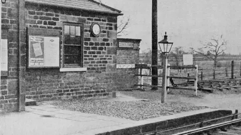 Black and white photo of Heighington Station from 1920. It is a brick building with a lampost outside and a sign which says "Gentlemen" on the right hand side of the building. The platform in front of the station is cobbled. 
