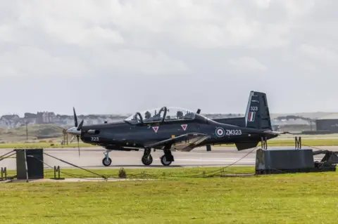 Getty Images A Beechcraft Texan T1 aircraft seen taxiing to runway to provide basic flying training at RAF Valley. The plane has two seats one in front of the other and is black