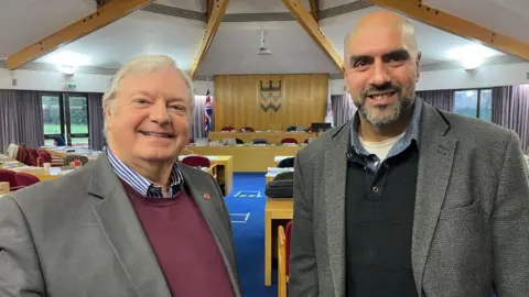 Two men smile towards the camera at the back of a council meeting room with blue carpet and rows of long tables facing towards two lines of desks at the front of the room. The seats are red. The man on the left is white haired and wears a maroon jumper, blue and white stripe shirt and grey jacket. The man on the right wears a flecked grey jacket over a jumper and shirt. He has a dark greying beard and shaved head.
