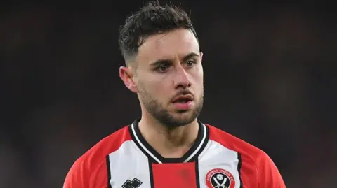 George Baldock playing for Sheffield United. He has dark hair with a beard and is wearing a red and white Sheffield United top. 