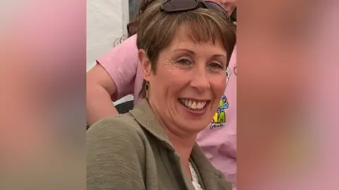 Pacemaker A woman has short length brown hair with sunglasses perched on top of her head. She is smiling, with a gold earring visible in her left ear. Behind her a child can be seen wearing a pink top.