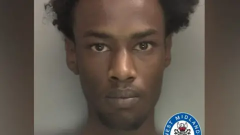 West Midlands Police A police mugshot showing a man with curly black hair and brown eyes, wearing a grey t-shirt. The West Midlands Police logo is in the bottom right-hand corner of the photo.