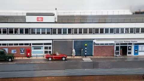A model railway station replicating the outside of Gloucester station. There is a model road, a zebra crossing, cars, a post box and people. It shows the entrance to the station with the words Gloucester Railway Station above the door. 