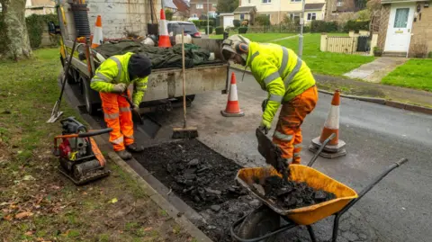 Two workers in high vis clothing are using spades to repair a pothole. There are beside a van with a wheelbarrow of tarmac