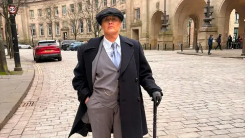 Woman dressed in three piece black and grey suit with a flat cap and cane.