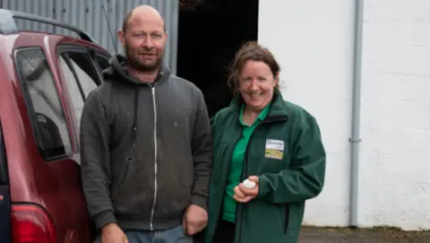 Family handout Shows a burgundy-coloured vehicle to the left then Vincent in blue jeans and a grey zip-up hooded to and Annie in a green T-shirt and darker green top, holding two eggs in her left hand