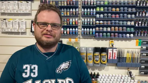 A man with short brown hair, moustache and beard, and wearing a blue American football top is standing in front of a wall of paint pots in a shop.