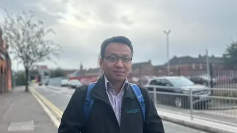 Colin Tang wearing a black coat with a purple and white stripped shirt beneath. He is wearing black glasses and has a blue backpack on his shoulders.