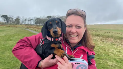 Kate Harrison is a young woman holding dachshund Bobbi in her arms. She is wearing a pink anorak and behind her is a long stretch of hillside with a path and a row of trees at the top