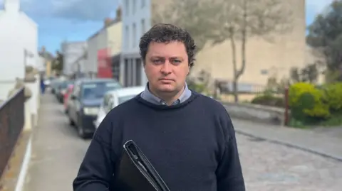 Alex Snowdon facing the camera, stood on Victoria street in Alderney. He is weaing a Navy Guernsey Jumper and holding a binder.