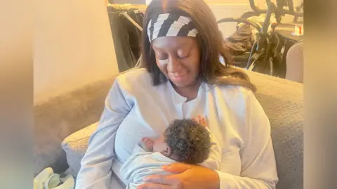 Modupe holds tiny Alpha against her chest and looks down and smiles at him. 