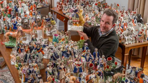 A man in a zip top with receding hair holds up a piece of Staffordshire pottery in the midst of a huge colourful collection of other pottery items