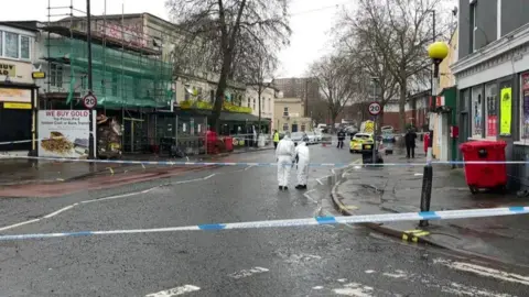 Two detectives in forensic suits look at the floor on a street behind police tape. There are a number of shops and a pub on the street, and it is a 