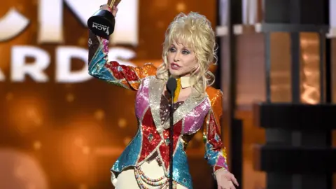 Getty Images/Ethan Miller Dolly holds up the award in the air. She wears a riding jacket type coloured shiny, sparkly jacket, with cream jodhpurs.