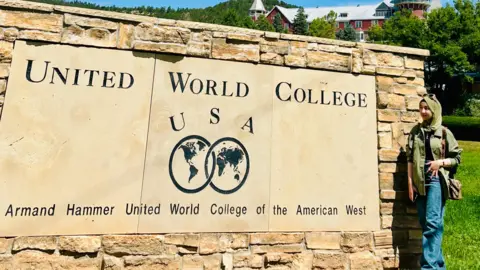 Aqdas Aqdas, a woman wearing a green jacket and blue jeans, posing in front of a United World College USA sign with the slogan "Armand Hammer United World College of the American West"