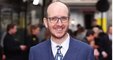 Getty Images Jack Thorne wears a blue suit and colourful tie and stands facing the camera with a broad smile