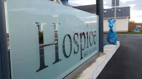 A glass sign that reads Hospice Isle of Man. A blue decorated statue of a wallaby can be seen next to the sign and a similarly painted dolphin statue is in the background.