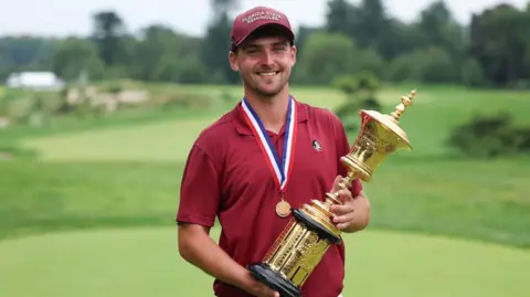 Whaley standing on a golf course holding a large gold trophy and wearing a medal around his neck. 