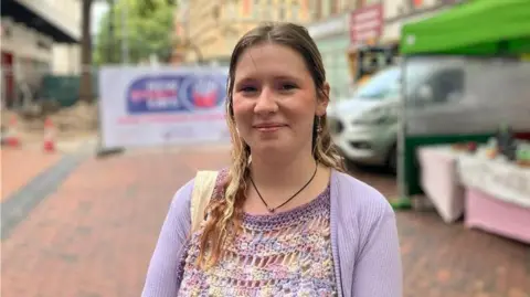 A young woman wearing a purple cardigan and crocheted top smiles, with a city street behind her. 