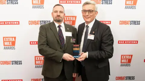 David is standing on the left and is wearing a dark green suit jacket, white shirt and blue and red tie. He has brown hair and is holding onto one side of a glass trophy. On the right is Admiral Sir Philip Jones who has silver white hair, and is wearing a white shirt, black suit jacket and black tie. He is holding on to the other side of the glass trophy.