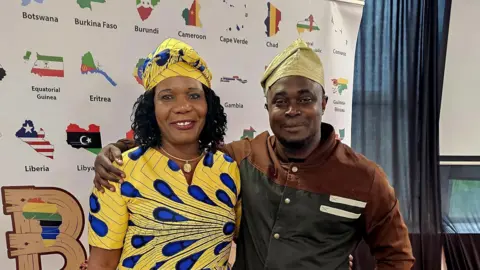 Aisha Iqbal/BBC A man and woman, both of African heritage, stand smiling. He has an arm around her shoulder. She wears a yellow and blue dress in a traditional style, and he wears a gold coloured cap. They stand in front of a display board bearing the names and flags/maps of several African countries.