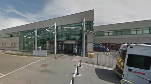 The front entrance to Ysbyty Gwynedd Hospital. Ambulance vans can be seen on the right hand side. A side reads: Main entrance" above automatic glass doors. A person walks towards the doors. 