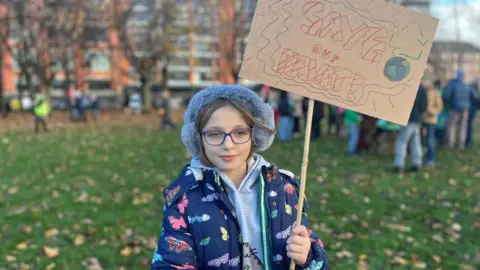 A child wearing a fur hat, glasses and a butterfly-patterned coat stands in a park holding a handmade cardboard sign that reads
